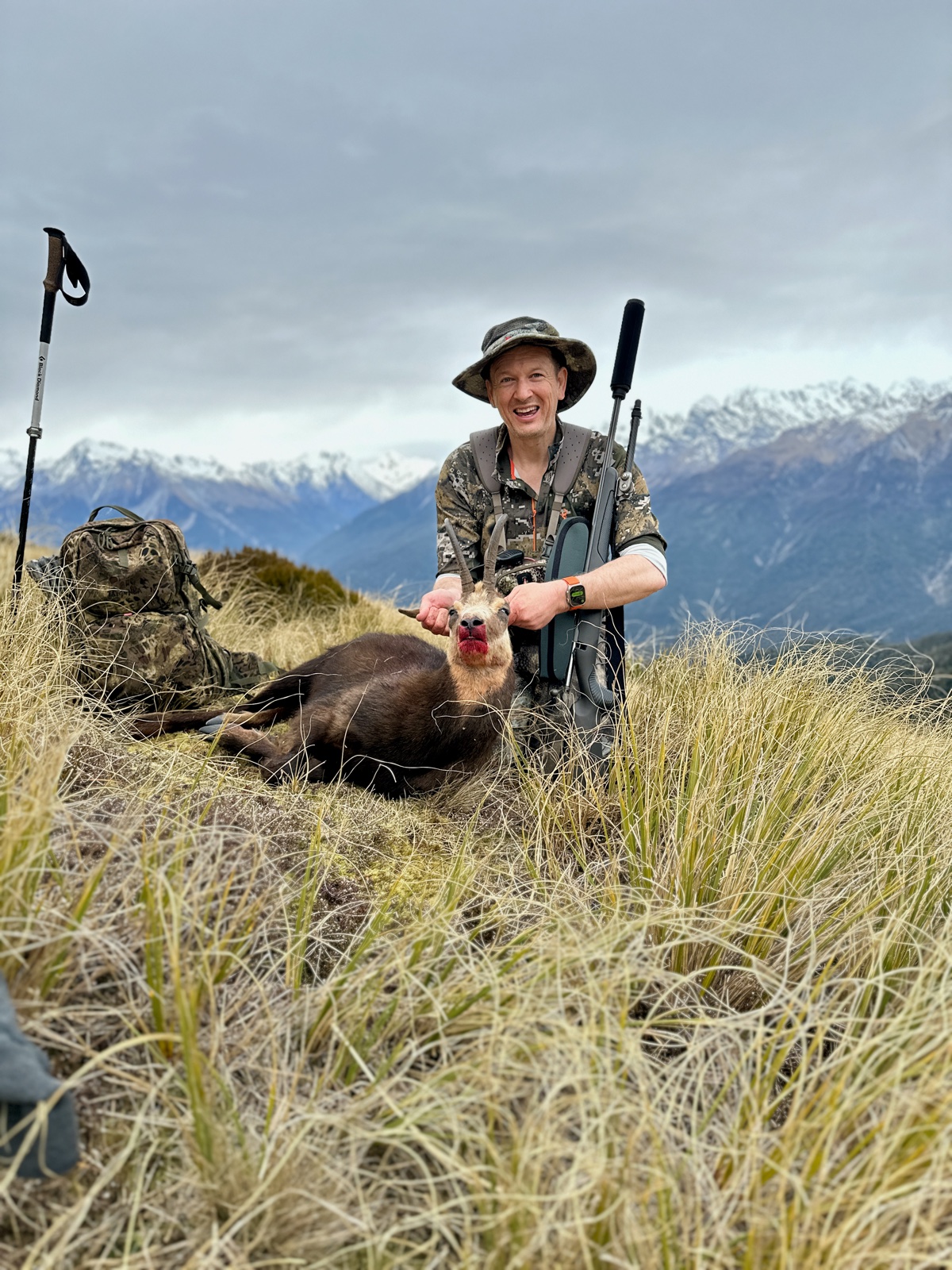 the developer kneeling in deep tussock with a chamois, a snow-covered range behind him under a grey sky.