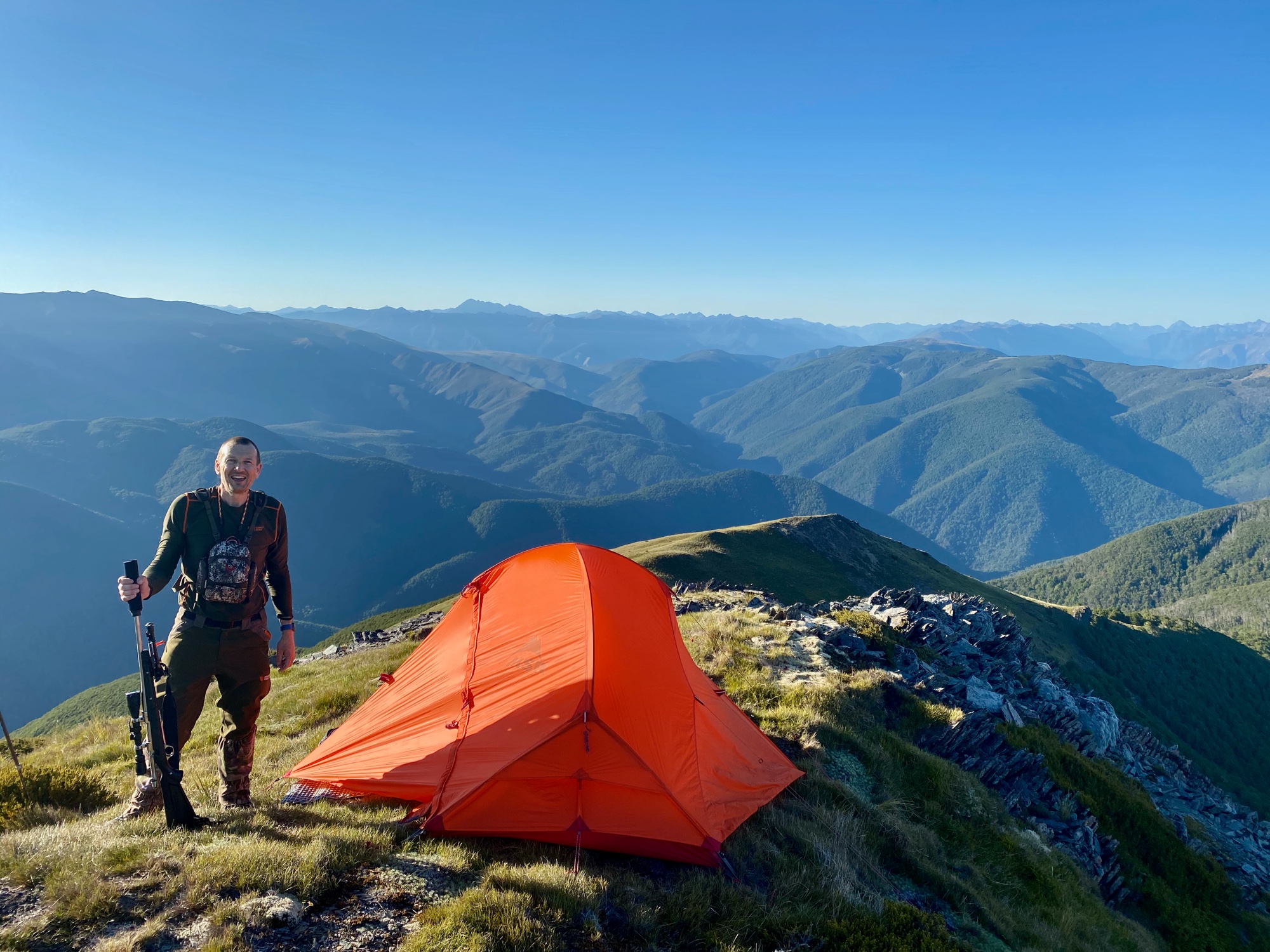 A camp on a tussock ridge at first light, with range after range of bush-covered valleys running away behind it.