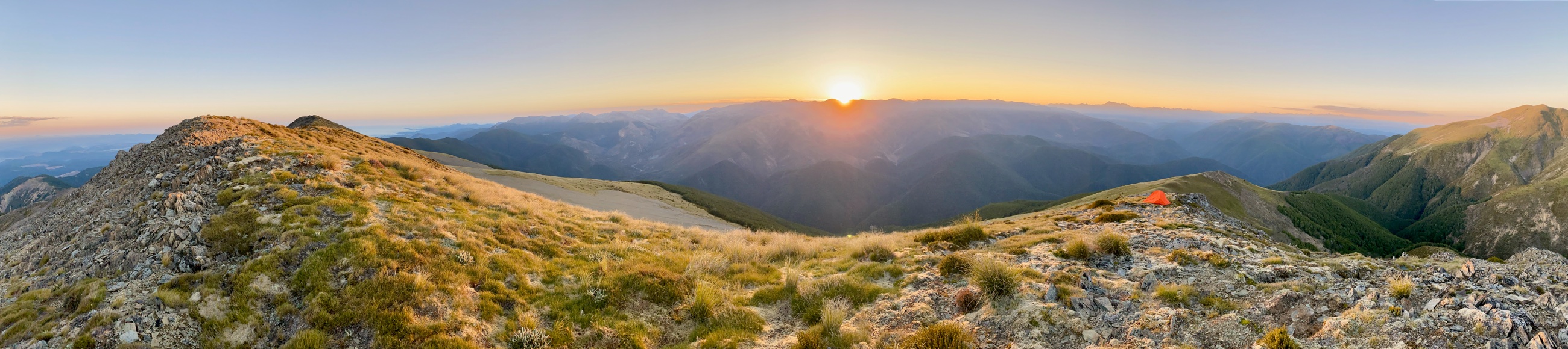 A wide panorama from a tops camp at sunrise, ranges receding into haze in every direction, a small orange tent on the ridge to the right.
